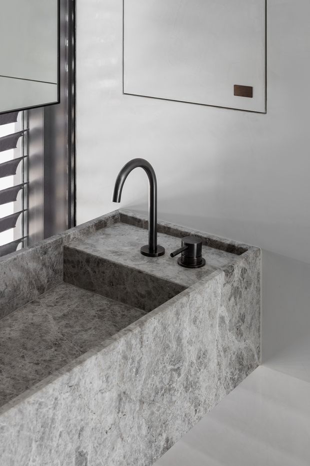 Close-up of a minimalist bathroom vanity with a substantial integrated grey-veined stone basin and benchtop. A matte black curved mixer tap and matching soap dispenser sit beside the basin. White walls, a black-framed louvre window, and a simple framed wall panel create a restrained monochromatic composition.