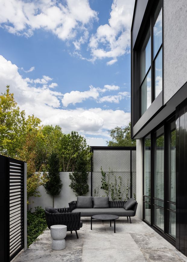 A contemporary outdoor courtyard lounge beside a modern two-storey building. The space features a concrete-paved terrace, black-framed full-height glazing and dark cladding, a grey outdoor sofa and armchair, a round coffee table, planted screening, and a translucent grid-pattern privacy panel. The image has a bright blue sky and leafy landscaping visible beyond the courtyard.