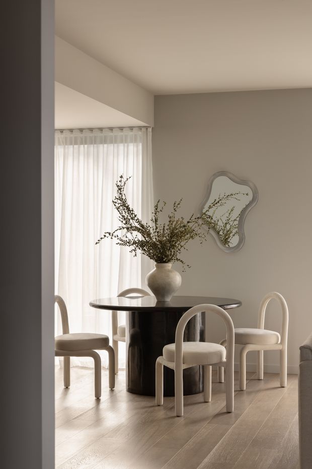 A refined dining area with a round black pedestal table, upholstered curved-back dining chairs, and a sculptural ceramic vase with branches as the centrepiece. Soft sheer curtains provide filtered daylight, while a wavy-edged wall mirror and neutral taupe walls create a calm, understated setting. Light timber flooring and minimalist architectural detailing complete the room.