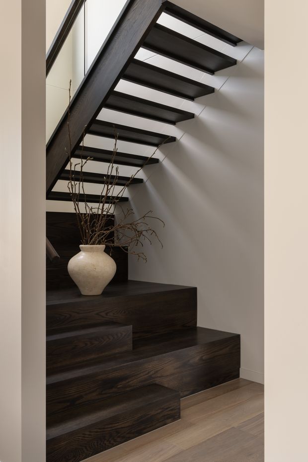 Interior view of a sculptural staircase with dark-stained timber treads, risers, and integrated stepped joinery. The open-riser stair rises along a white wall beneath dark exposed structural beams, with a large neutral ceramic vase and branches placed on the steps. A pale timber floor contrasts with the dark stair detailing.