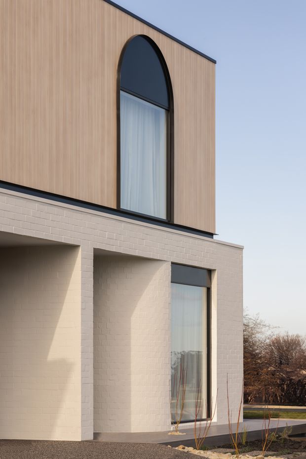 A modern two-storey residential facade with a pale rendered brick lower level and warm vertical timber-look cladding above. The facade features tall dark-framed glazing, including a prominent arched upper window and a full-height lower window, with clean geometric projections and a restrained contemporary palette. Minimal landscaping and a paved entry edge are visible in the foreground.