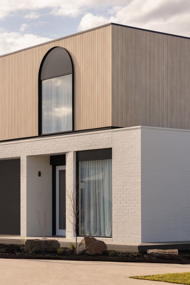 Exterior view of a contemporary two-storey residence with a clean geometric form. The lower level is clad in white-painted brick, while the upper level features vertically ribbed timber-toned cladding. A tall arched window with dark framing is a prominent feature, complemented by large ground-floor glazing, a recessed entry, dark trim, and minimalist landscaping with large rocks.