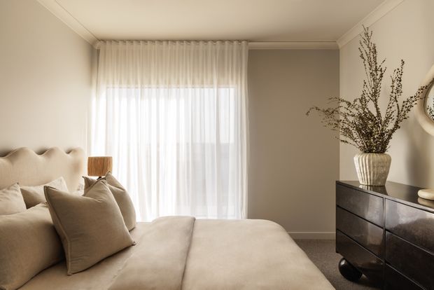 A softly styled bedroom in warm neutral tones, featuring a plush upholstered bed with layered beige pillows, full-height sheer curtains over a large window, a dark timber or stained-wood dresser, and a sculptural ceramic vase with tall dried branches. The room has subtle wall panelling or trim, a simple tray ceiling detail, and gentle natural light, creating a calm, refined atmosphere.