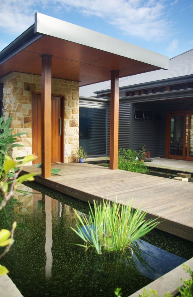 Exterior view of a contemporary courtyard entry featuring a timber deck and covered porch supported by timber posts, a natural stone-clad wall with timber entry door, dark corrugated metal cladding, and a reflecting pond with planted landscaping.