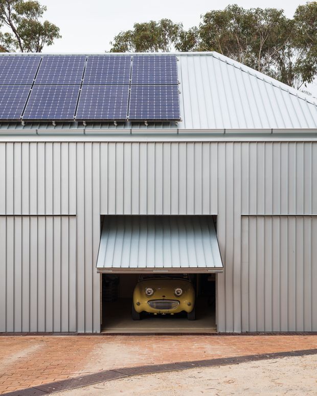 Exterior view of a corrugated metal-clad garage or outbuilding with a pitched metal roof, rooftop solar panels, and a central roller-style garage opening with a small awning. A vintage yellow car is visible inside. The building sits beside a brick-paved driveway or forecourt, with trees in the background.