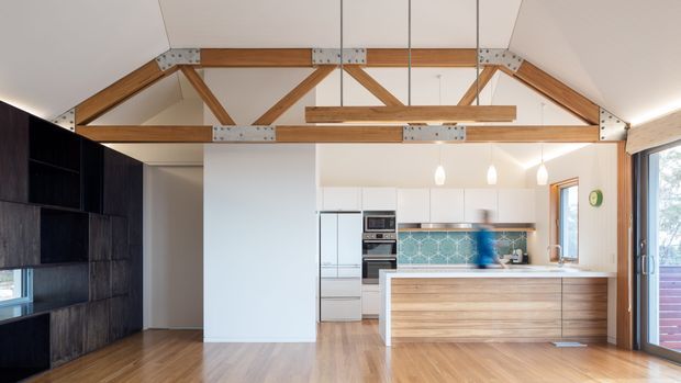 Bright open-plan kitchen and living area with a prominent exposed timber truss ceiling. The kitchen features white cabinetry, a timber island and benchtop, and a blue patterned tile splashback. Dark built-in joinery lines the left wall, while glazed doors on the right open toward an outdoor area.
