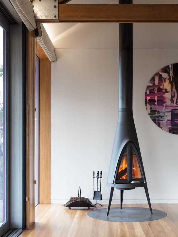 Interior view of a warm, minimalist living area featuring a freestanding black suspended wood-burning fireplace with a visible flame, set on a circular hearth mat. The room has pale timber flooring, white walls, tall timber-framed glazing and exposed timber structural beams, with fire tools beside the fireplace and a partially visible abstract artwork.