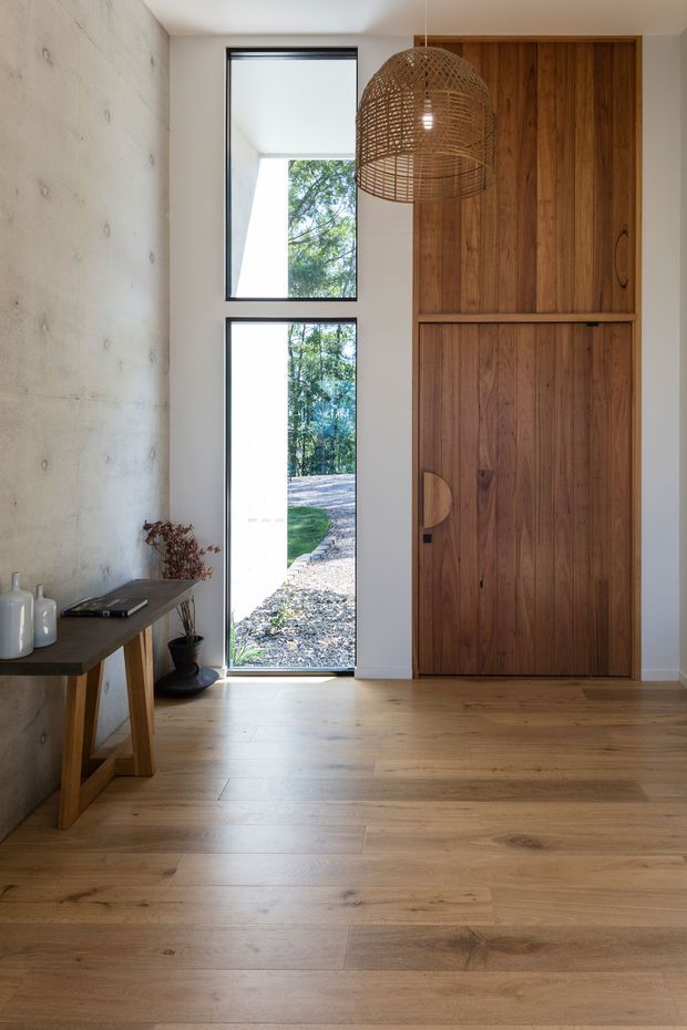 A minimalist residential entryway with a full-height timber pivot door and matching built-in timber paneling, pale timber flooring, a raw concrete feature wall, and narrow black-framed glazing. A woven pendant light hangs above a simple console table, with greenery visible outside.