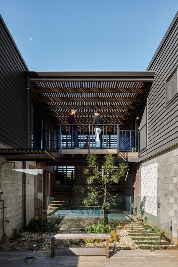 A contemporary multi-level courtyard exterior with dark horizontal cladding, concrete block walls, timber decking and a glass-edged water feature. A raised balcony and pedestrian bridge sit beneath a timber slatted pergola, connecting the surrounding building wings. The courtyard includes gravel landscaping, stepping stones, a small tree and a timber bench, with strong natural daylight and a clear blue sky.
