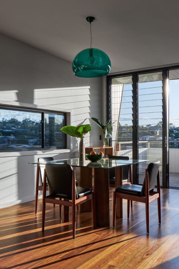 Sunlit dining area with a rectangular glass-top dining table and four dark timber dining chairs. The room features warm timber flooring, white horizontal wall cladding, extensive black-framed louvred glazing opening to a balcony, and a distinctive teal pendant light. Indoor plants and strong afternoon shadows add a warm, relaxed character.