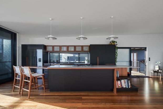 A spacious contemporary open-plan kitchen and dining area with a large dark island, timber breakfast stools, integrated cabinetry, and a long horizontal window above the benchtop. Three suspended pendant lights hang over the island, while warm timber flooring and timber shelving add contrast to the black and white joinery. A hallway or living area is visible beyond the kitchen.