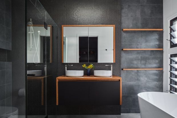 A stylish contemporary bathroom featuring a double vanity with white vessel basins, dark timber-framed cabinetry, a large timber-framed mirror, black small-format textured wall tiling, grey large-format wall tiles, open timber shelving, a glass shower enclosure, and a partially visible white freestanding bathtub. White louvred window shutters provide daylight at the right.