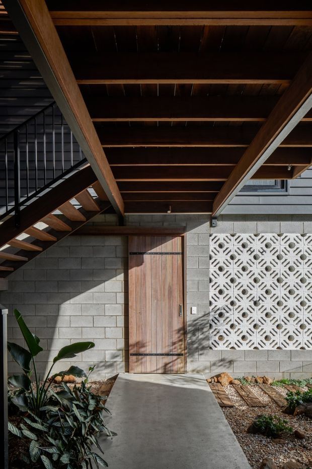 A covered exterior entry courtyard beneath a deep timber-beamed roof. The facade combines grey concrete block walls, dark horizontal cladding, a vertical timber door with black strap hinges, and a decorative white breeze-block screen. A black metal stair with timber treads runs along the left side, while a concrete path and planted gravel garden lead to the entrance. Strong sunlight creates geometric shadows across the facade and walkway.