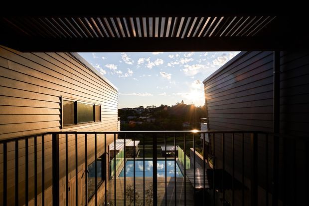 A contemporary architectural exterior view from an elevated balcony or terrace between two horizontally clad building volumes. Dark metal balustrades and a slatted overhead screen frame a swimming pool below, with warm low-angle sunlight and a suburban landscape beyond.