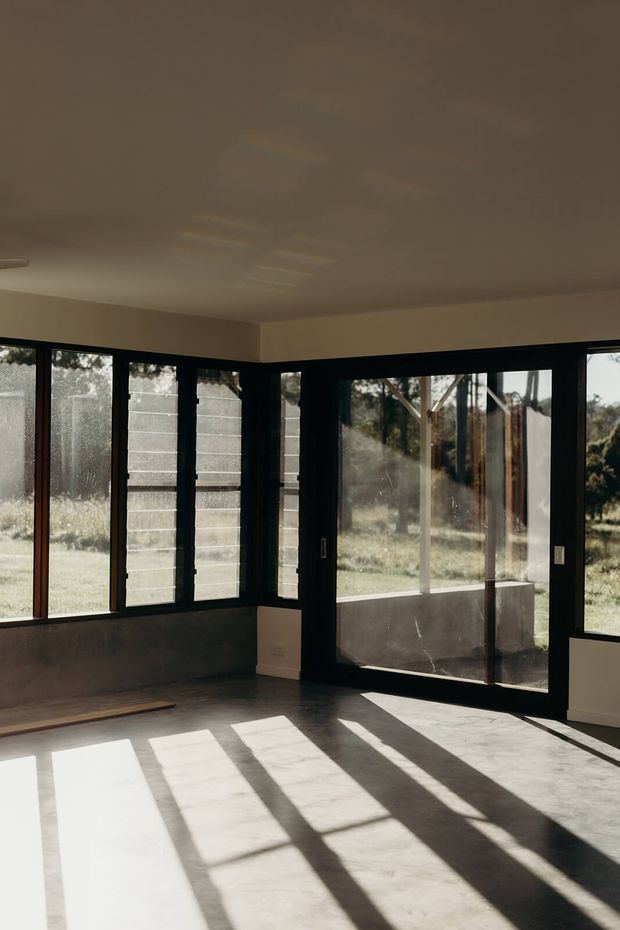 An empty, sunlit interior room with expansive black-framed windows and glazed doors wrapping around two walls. Strong afternoon light casts long geometric shadows across the pale concrete or stone-like floor. The room has plain light walls, a low flat ceiling, and a view toward a grassy, wooded landscape outside.