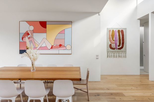 Bright contemporary dining area with a long warm timber dining table and light upholstered chairs, set against white walls and pale timber flooring. Abstract geometric artwork and a woven wall hanging add colourful accents, while an open hallway or adjoining space is visible to the right.