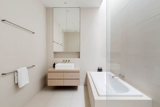 Bright, minimalist bathroom with a freestanding-style white bathtub enclosed by a glass shower screen, a wall-mounted timber vanity with white basin, mirrored cabinetry, pale tiled surfaces, and simple metal towel rails.