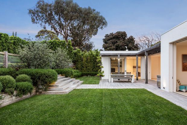 A landscaped residential courtyard with a broad manicured lawn, layered garden beds, mature trees and clipped shrubs. Concrete steps lead to a pale timber deck and covered outdoor entertaining area beside a low, flat-roofed contemporary building. The space includes an outdoor dining table and benches, with warm interior light visible through the glazing.