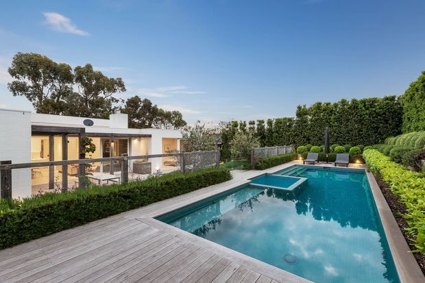 A landscaped modern residential property viewed from an elevated outdoor deck, featuring a large rectangular swimming pool with an integrated raised spa, timber decking, manicured hedges, sculpted shrubs, and a low flat-roof white house with extensive glazing and a sheltered outdoor living area.