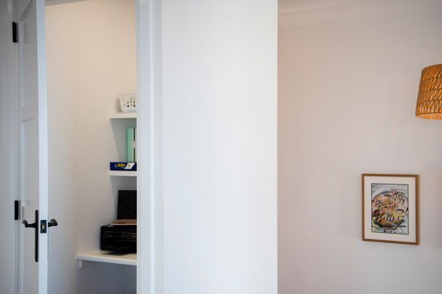 A bright interior view showing a white-painted hallway or room corner with an open door leading to a compact built-in home office nook. The nook includes white shelving, a desk surface, and office equipment. A framed artwork and woven pendant light add warm decorative accents to the adjacent wall.