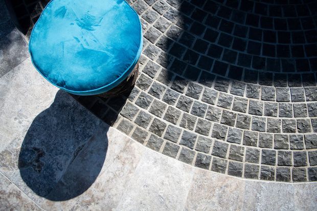 Close-up overhead view of a curved paved surface featuring concentric bands of small dark stone or brick pavers set into a pale stone or concrete ground. A bright turquoise circular object sits at the upper left, casting a strong shadow. The image emphasizes geometric paving patterns, texture, and high-contrast sunlight.