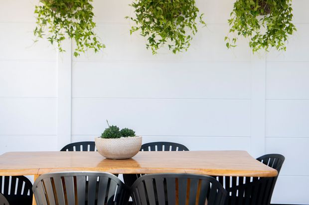 Bright dining area with a long light timber dining table, black slatted dining chairs, a textured stone planter centerpiece, and hanging trailing greenery against a white horizontally panelled wall.