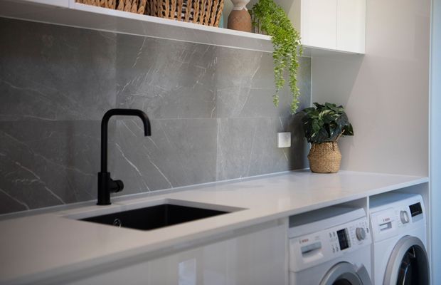 A contemporary laundry with a long white engineered-stone benchtop, integrated dark sink and matte black gooseneck tap. The wall features a large-format grey stone-look splashback, with glossy white cabinetry and front-loading white washing and drying machines below. Open shelving, woven storage, and potted greenery add warmth.
