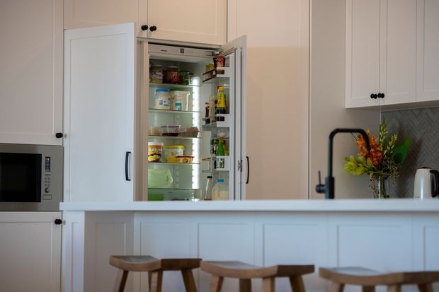 Bright traditional-style kitchen with white raised-panel cabinetry, a large open refrigerator integrated into the joinery, a pale island or peninsula with decorative panel detailing, and timber stools. A microwave is built into the cabinetry, while a patterned tile splashback, black hardware, and a vase of flowers add contrast.