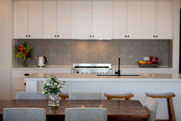 A bright, refined open-plan kitchen and dining area. The kitchen features extensive white shaker-style cabinetry, a large white island with integrated sink and dark tapware, a grey herringbone-patterned splashback, and a stainless-steel rangehood and cooktop. In the foreground, a timber dining table is paired with upholstered dining chairs and stools, with a floral arrangement as a centrepiece.
