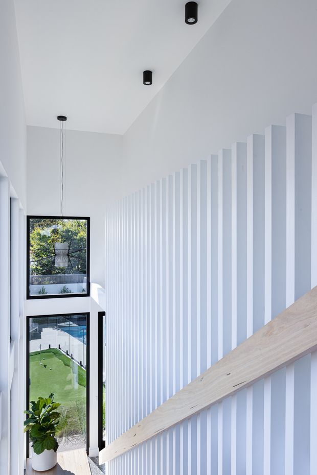 A bright, minimalist stairway with a pale timber handrail running alongside a dramatic wall of closely spaced white vertical battens. Black cylindrical ceiling lights and tall windows bring in natural light and views of greenery and a backyard below.