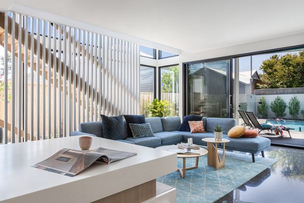Bright contemporary open-plan living room with a low blue sectional sofa, white sculptural coffee tables, and a large white island or console in the foreground. Floor-to-ceiling glazing opens toward a pool courtyard, while a vertical timber slat screen forms a striking feature partition. The space has polished reflective flooring, a neutral palette, and abundant natural light.