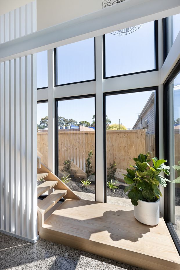 Bright contemporary interior showing a timber stair and built-in window seat beside full-height black-framed glazing. White vertical slats form a balustrade or screen, while sunlight falls across a polished exposed-aggregate floor and pale timber joinery. A small planted courtyard is visible through the windows.