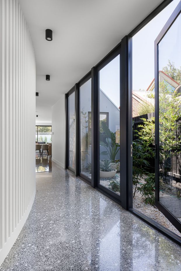 A bright contemporary interior corridor lined with full-height black-framed glazing on one side, opening onto a planted courtyard. The opposite wall has vertical white ribbed panelling, while the polished terrazzo floor reflects daylight and compact black ceiling spotlights. A dining area is visible at the far end.
