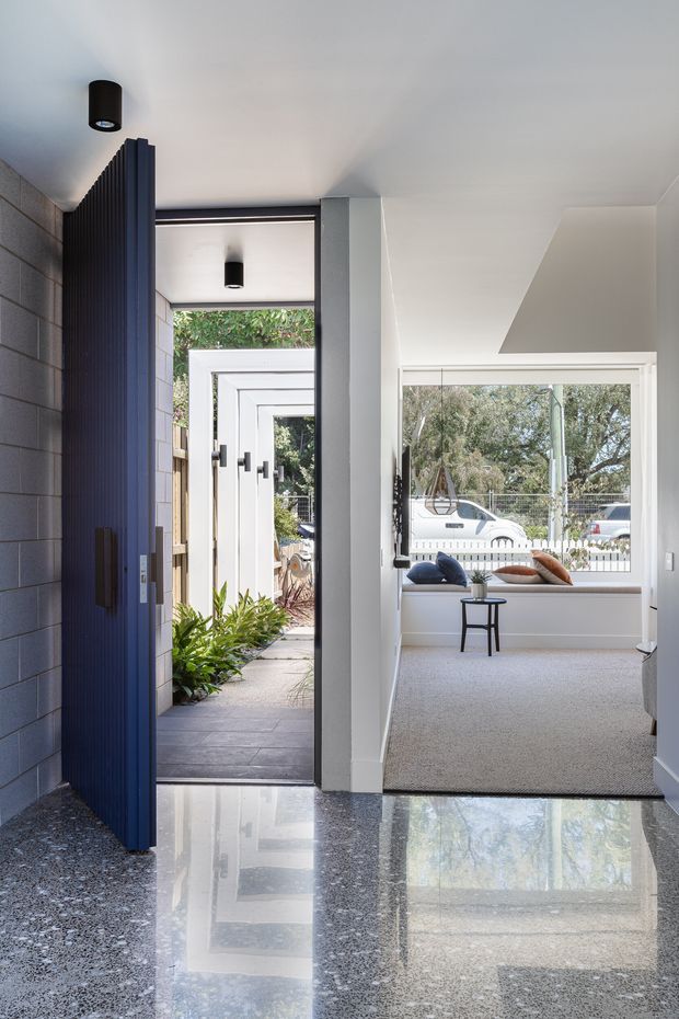A bright contemporary entry hall with a dramatic dark vertical-slat pivot door, polished terrazzo flooring, pale walls, and a direct view through to a carpeted sitting area with a built-in window seat. The entry opens onto a landscaped courtyard, while a large window brings in views of trees and the street beyond. Minimal black ceiling lights and dark hardware provide contrast.
