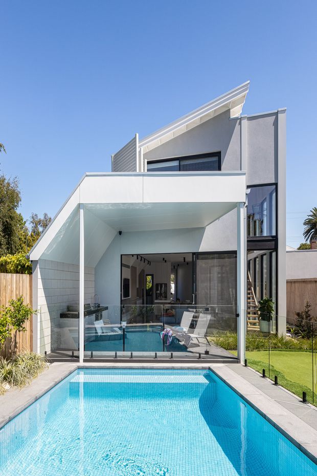 Bright exterior view of a contemporary multi-level home with a sharply angled pitched roof, white rendered walls, dark-framed glazing, and a covered outdoor entertaining area. A long tiled swimming pool occupies the foreground, with frameless glass pool fencing, a compact lawn, timber boundary fencing, and visible indoor-outdoor living spaces beyond.