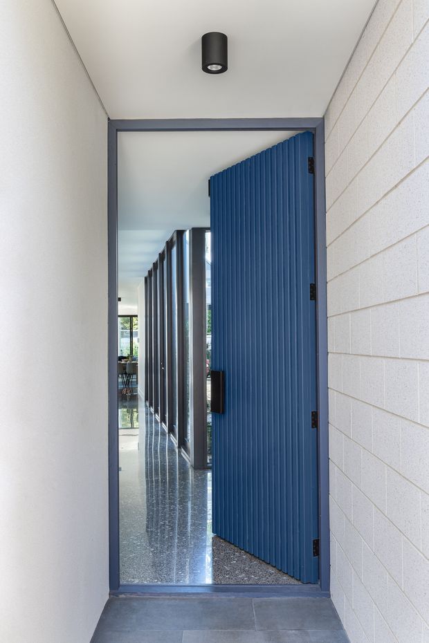 A narrow entry passage with a blue vertically ribbed pivot door, dark grey metal door frame, pale painted walls, and a white painted masonry block wall. The open doorway reveals a bright modern interior beyond with reflective polished flooring, glazed partitions, and additional dark-framed doors. A simple black cylindrical ceiling light provides illumination.