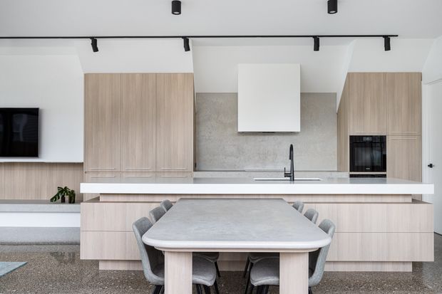 Contemporary open-plan kitchen and dining area with pale timber cabinetry, a large central island, integrated appliances, concrete-look splashback, and a light stone-look benchtop. A dining table with upholstered chairs sits in the foreground, while black track lighting and a wall-mounted television add modern detailing.