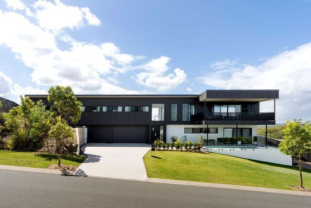 A contemporary multi-storey residence viewed from the street, featuring a long low-profile form, dark vertical cladding, extensive glazing, a projecting upper-level volume, and a broad landscaped front lawn. The property includes an integrated double garage and a paved driveway, with distant hills visible beyond the site.