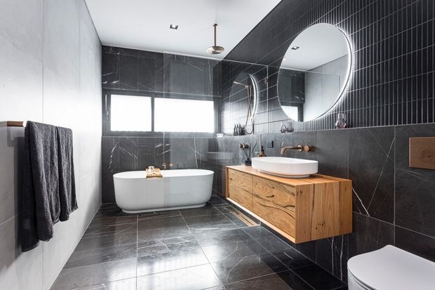 Contemporary bathroom featuring dark marble-look wall and floor tiles, a freestanding white bathtub beneath a wide window, a floating timber vanity with a white vessel basin, wall-mounted brass tapware, a round illuminated mirror, black vertical wall panelling, and a partially visible toilet. The space has a refined, high-contrast monochromatic palette with warm timber and brass accents.