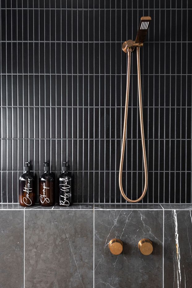Close-up of a refined shower wall featuring slim black vertical tiles with horizontal grout bands, a dark grey marble-look tiled lower wall, and a copper/bronze handheld shower with matching controls. Three dark amber bath product bottles sit on the tiled ledge. The composition is minimalist, dark, and luxurious.