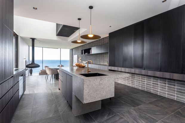 A sophisticated modern kitchen with a large concrete island, dark vertically ribbed cabinetry, grey tiled flooring, and a horizontal grey tiled splashback. Brass-toned tapware and warm pendant lights provide subtle highlights. The open-plan space extends to a dining area and floor-to-ceiling glazing with a distant ocean view.