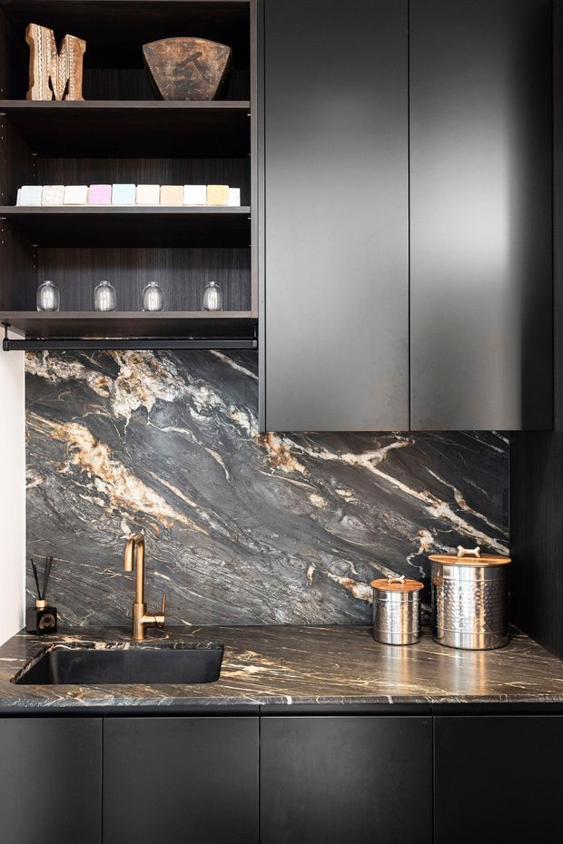 A compact, highly refined kitchen or kitchenette featuring matte charcoal-black cabinetry, open dark timber shelving, and dramatic veined stone applied across the splashback and benchtop. A deep black undermount sink with a brushed brass tap sits below the shelving, while metallic canisters and curated objects add contrast. The palette is dark, luxurious, and strongly material-focused.