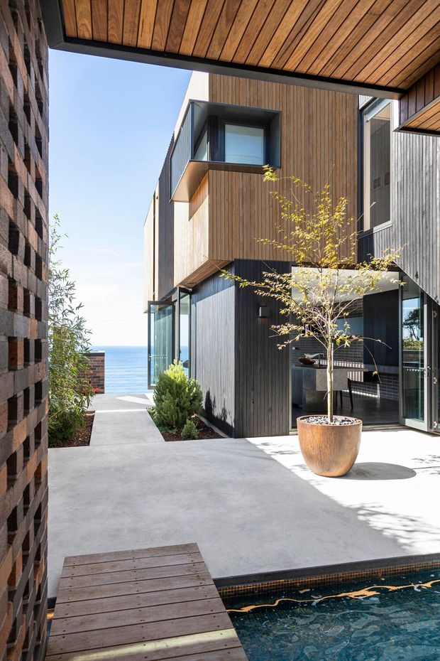 A bright contemporary courtyard between two dark brick and timber-clad building volumes, with large glazed openings, a timber-lined overhead soffit, concrete paving, and a small tiled swimming pool in the foreground. A timber deck bridge, potted tree, and coastal ocean view beyond create a refined indoor-outdoor setting.