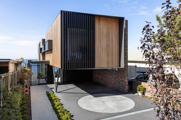 Contemporary two-storey residential exterior on a sloping suburban site, featuring a projecting upper volume clad in warm vertical timber and screened with black vertical fins. The lower level combines dark brickwork, black metal framing, a covered carport, and a broad dark paved driveway with a circular patterned feature. Timber fencing, neighbouring buildings, and planted edges frame the approach.