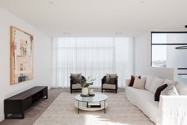 Bright, refined contemporary living room with a large white sectional sofa, two dark-framed lounge chairs, a round glass coffee table, and a low dark timber media console. Sheer floor-to-ceiling curtains diffuse abundant daylight, while a large abstract artwork and neutral textured rug add understated visual interest. Black-framed internal glazing or a window opening is visible at right.