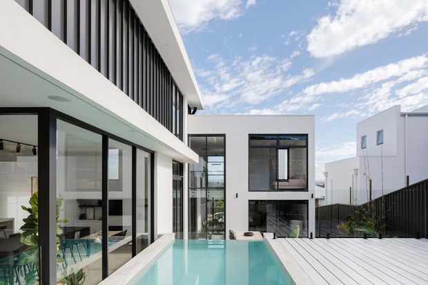 Bright contemporary multi-level residential courtyard viewed from an outdoor pool terrace. White rendered cubic forms, extensive black-framed glazing, vertical facade screening, and a pale deck surround a narrow turquoise swimming pool. Interior living and dining areas are visible through the glazing, with a clear urban residential setting and blue sky beyond.