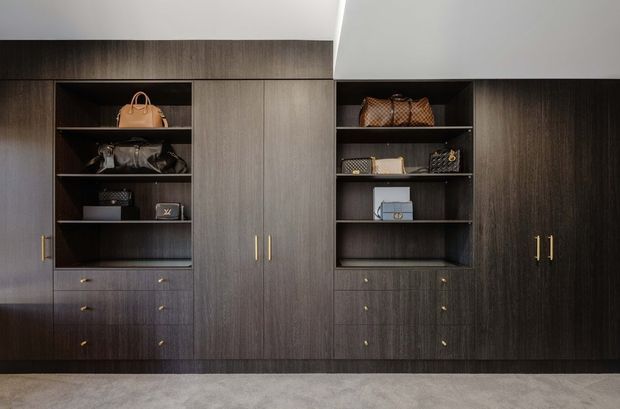 A wall-to-wall built-in wardrobe and display storage installation with dark wood-grain cabinetry, open shelving, drawers, and brass-toned handles. Handbags and accessories are arranged on the shelves, while a light neutral carpet and white ceiling provide contrast. The composition is symmetrical and refined, with clean contemporary joinery.