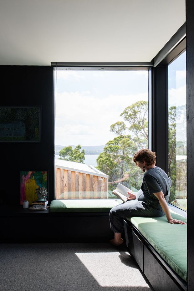 A compact, moody reading nook or study with black walls, charcoal carpet, dark-framed corner glazing, and built-in window seating upholstered in muted sage green. A person is seated reading beside a small display ledge with artwork and books. The large windows provide bright natural light and views toward trees, water, and neighbouring rooftops.