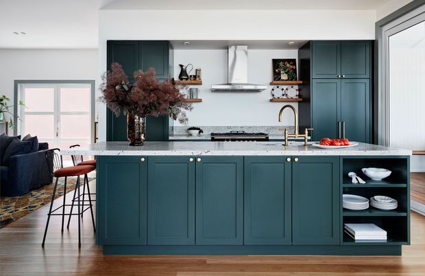 A refined contemporary kitchen with a large dark teal island featuring framed cabinetry and brass hardware, a pale stone benchtop, open shelving, and a white tiled backsplash. Dark teal full-height cabinetry surrounds a stainless-steel rangehood and cooktop. Timber flooring, black-framed glazing, bar stools, and a partially visible adjoining living area complete the scene.