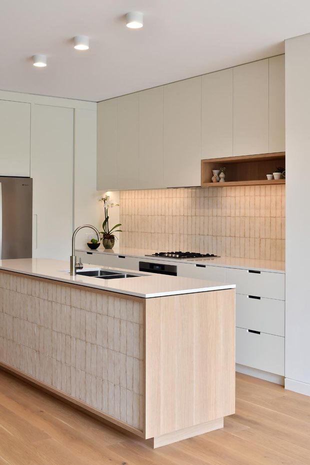 Bright contemporary kitchen featuring a large island with an integrated sink, pale cabinetry, a light timber island end panel, vertically textured beige tile cladding on the island and splashback, timber flooring, and a recessed timber display shelf. Minimal recessed ceiling lights and stainless-steel appliances complete the refined neutral scheme.